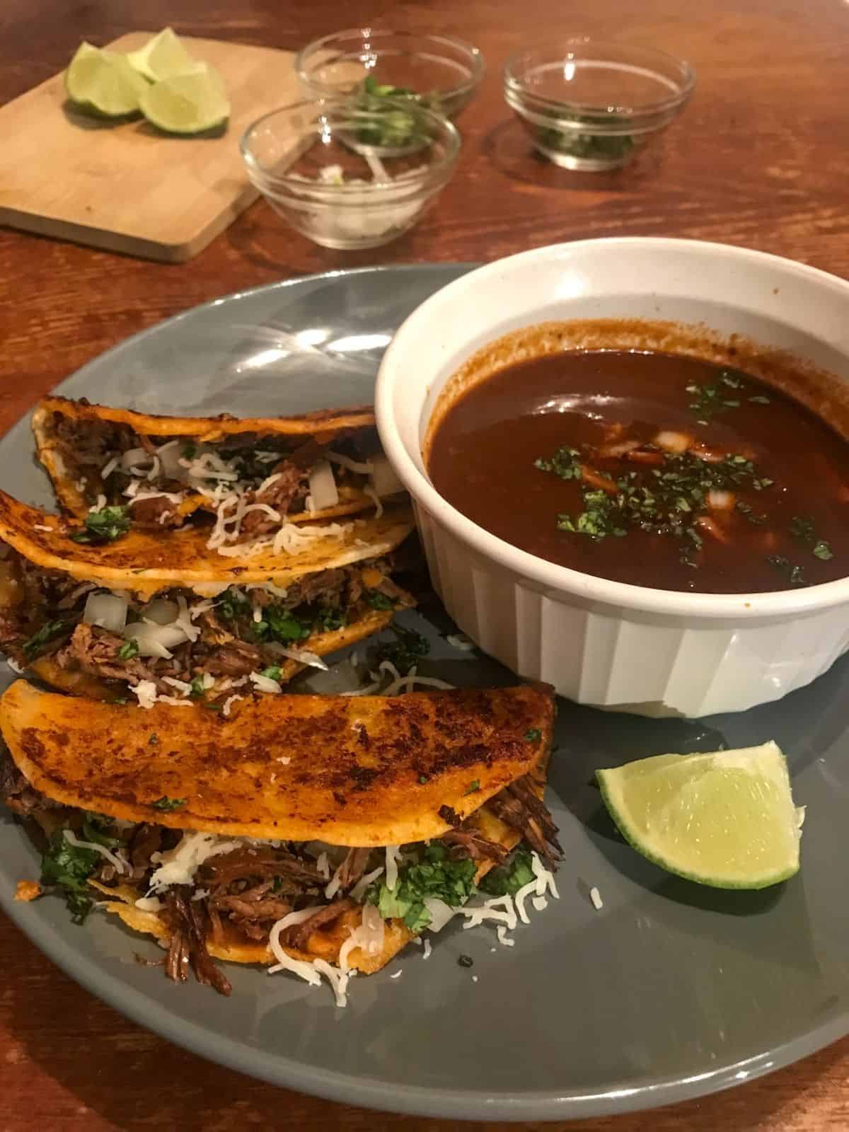 birria quesa tacos with consome with toppings in the background