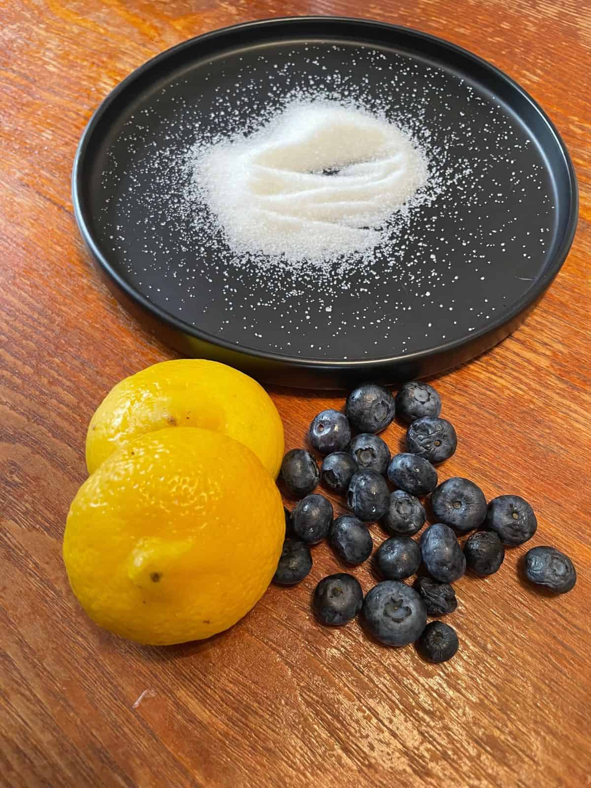 overhead shot of lemon halves, blueberries, and sugar on a black plate